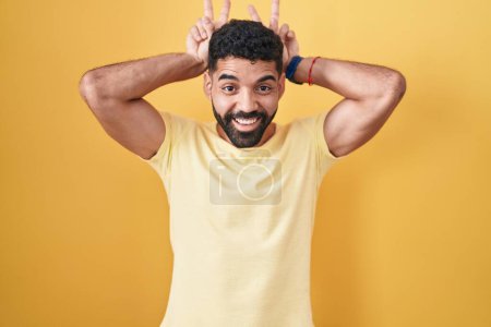 Foto de Hombre hispano con barba de pie sobre fondo amarillo posando divertido y loco con los dedos en la cabeza como orejas de conejo, sonriendo alegre - Imagen libre de derechos