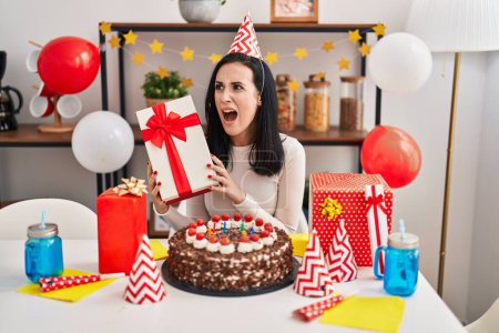 Photo for Hispanic woman celebrating birthday with cake holding present angry and mad screaming frustrated and furious, shouting with anger. rage and aggressive concept. - Royalty Free Image