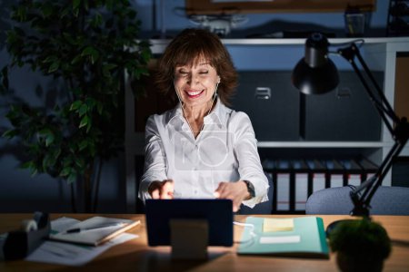 Téléchargez les photos : Femme d'âge moyen travaillant au bureau la nuit pointant vers vous et la caméra avec les doigts, souriant positif et joyeux - en image libre de droit