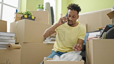 Photo for African american man unpacking cardboard box talking on smartphone looking upset at new home - Royalty Free Image