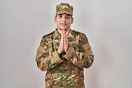 Téléchargez les photos : Jeune homme arabe portant l'uniforme de l'armée de camouflage priant avec les mains ensemble demandant pardon souriant confiant. - en image libre de droit