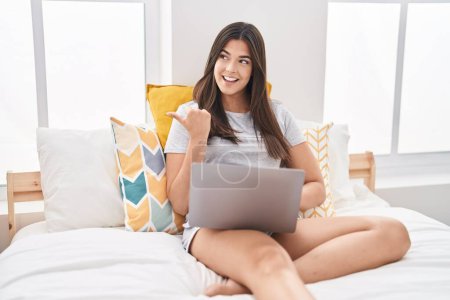 Téléchargez les photos : Hispanique femme utilisant ordinateur portable sur le lit pointant pouce vers le côté souriant heureux avec bouche ouverte - en image libre de droit