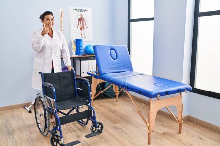 Téléchargez les photos : Femme hispanique d'âge moyen avec fauteuil roulant à la clinique de physiothérapie souriant heureux et positif, pouce levé faire excellent et signe d'approbation - en image libre de droit