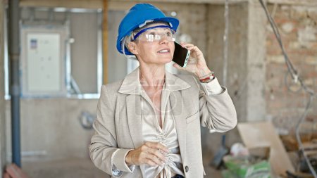 Téléchargez les photos : Moyen Age femme blonde architecte souriant confiant parler sur smartphone sur le chantier de construction - en image libre de droit
