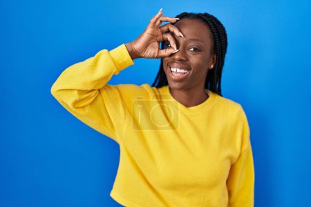 Photo for Beautiful black woman standing over blue background doing ok gesture with hand smiling, eye looking through fingers with happy face. - Royalty Free Image