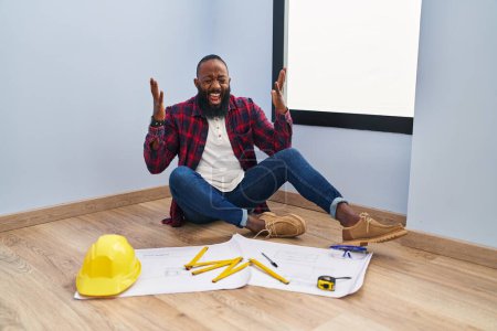 Téléchargez les photos : Homme afro-américain assis sur le sol à la nouvelle maison en regardant les plans célébrant fou et fou de succès avec les bras levés et les yeux fermés hurlant excités. concept gagnant - en image libre de droit