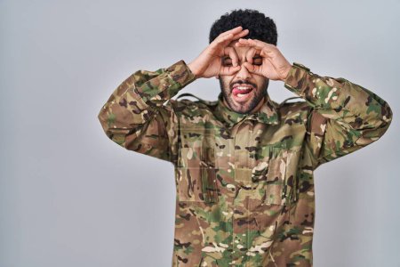 Téléchargez les photos : Homme arabe portant un uniforme de camouflage de l'armée faisant un geste correct comme des jumelles sortant la langue, les yeux regardant à travers les doigts. expression folle. - en image libre de droit