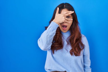 Photo for Hispanic young woman standing over blue background peeking in shock covering face and eyes with hand, looking through fingers with embarrassed expression. - Royalty Free Image