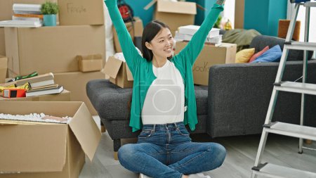 Foto de Joven mujer china sonriendo confiada sentada en el suelo estirando los brazos en un nuevo hogar - Imagen libre de derechos