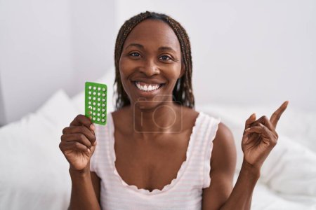 Téléchargez les photos : Femme afro-américaine tenant des pilules contraceptives souriant heureux pointant avec la main et le doigt vers le côté - en image libre de droit