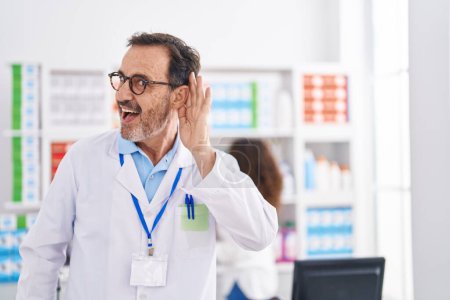 Photo for Middle age hispanic man working at pharmacy drugstore smiling with hand over ear listening and hearing to rumor or gossip. deafness concept. - Royalty Free Image