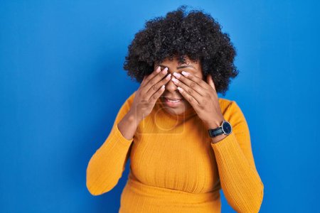 Téléchargez les photos : Femme noire avec les cheveux bouclés debout sur fond bleu frottant les yeux pour la fatigue et les maux de tête, somnolent et l'expression fatiguée. problème de vision - en image libre de droit
