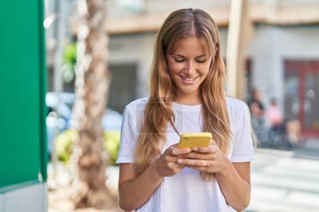 Téléchargez les photos : Jeune fille blonde souriant confiant en utilisant smartphone à la rue - en image libre de droit
