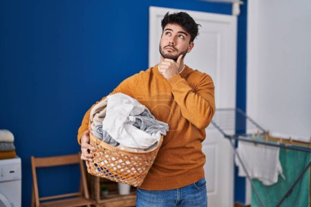 Photo for Hispanic man with beard holding laundry basket at laundry room serious face thinking about question with hand on chin, thoughtful about confusing idea - Royalty Free Image