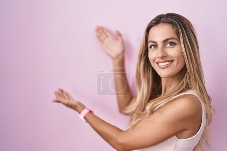 Foto de Mujer rubia joven de pie sobre fondo rosa invitando a entrar sonriente natural con la mano abierta - Imagen libre de derechos