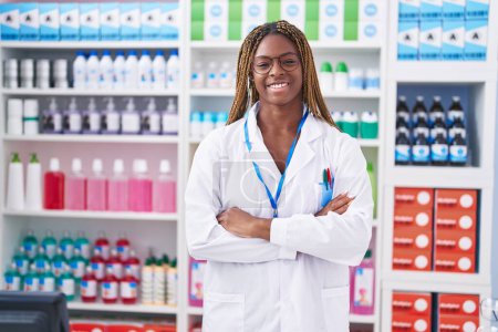 Foto de Farmacéutica afroamericana sonriendo confiada de pie con los brazos cruzados gesto en la farmacia - Imagen libre de derechos
