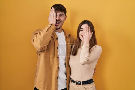 Téléchargez les photos : Jeune couple hispanique debout sur fond jaune couvrant un œil avec la main, sourire confiant sur le visage et émotion surprise. - en image libre de droit