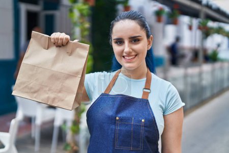 Photo for Young girl with blue hair wearing professional waitress apron holding delivery paper bag looking positive and happy standing and smiling with a confident smile showing teeth - Royalty Free Image