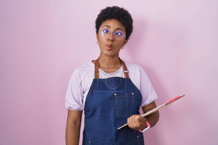 Foto de Mujer afroamericana joven con delantal de camarera profesional sujetando portapapeles haciendo cara de pez con labios, gesto loco y cómico. expresión divertida. - Imagen libre de derechos