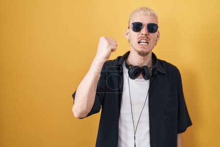 Photo for Young caucasian man wearing sunglasses standing over yellow background angry and mad raising fist frustrated and furious while shouting with anger. rage and aggressive concept. - Royalty Free Image