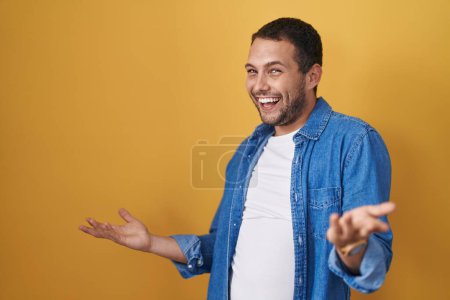 Foto de Hombre hispano de pie sobre fondo amarillo sonriendo alegre con los brazos abiertos como bienvenida amistosa, saludos positivos y confiados - Imagen libre de derechos
