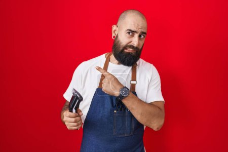 Téléchargez les photos : Jeune homme hispanique avec barbe et tatouages portant tablier de barbier tenant rasoir pointant de côté inquiet et nerveux avec index, expression inquiète et surprise - en image libre de droit