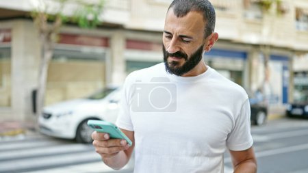 Téléchargez les photos : Jeune homme hispanique utilisant un smartphone avec un visage sérieux à la rue - en image libre de droit