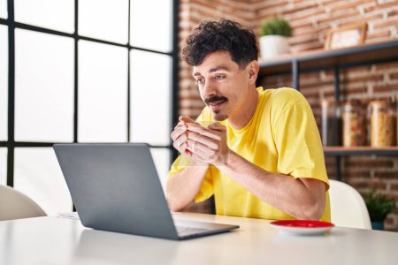 Téléchargez les photos : Young caucasian man using laptop drinking coffee at home - en image libre de droit