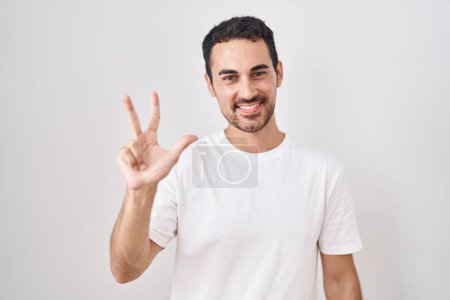 Foto de Hombre hispano guapo de pie sobre fondo blanco mostrando y señalando hacia arriba con los dedos número tres mientras sonríe confiado y feliz. - Imagen libre de derechos