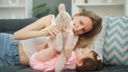 Photo for Mother and daughter lying on sofa playing with teddy bear at home - Royalty Free Image
