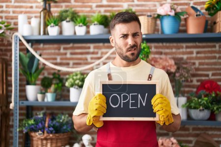 Photo for Handsome hispanic man working at florist holding open sign clueless and confused expression. doubt concept. - Royalty Free Image