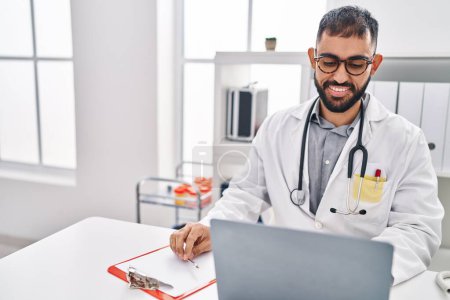 Photo for Young hispanic man doctor using laptop writing medical report at clinic - Royalty Free Image