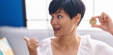 Foto de Joven mujer asiática con pelo corto sosteniendo moneda criptomoneda tron apuntando pulgar hacia el lado sonriendo feliz con la boca abierta - Imagen libre de derechos