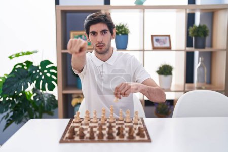 Foto de Joven hispano jugando al ajedrez sentado en la mesa señalando con el dedo a la cámara y a ti, gesto confiado y serio - Imagen libre de derechos