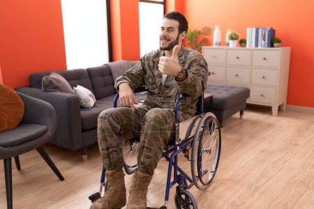 Photo for Young hispanic soldier man injured sitting on wheelchair smiling happy and positive, thumb up doing excellent and approval sign - Royalty Free Image