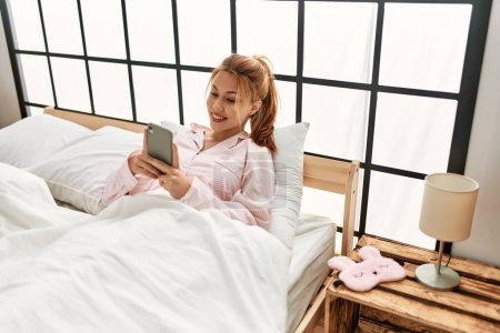 Téléchargez les photos : Jeune femme caucasienne utilisant smartphone assis sur le lit dans la chambre - en image libre de droit