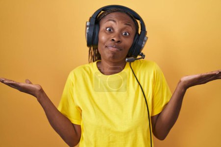Foto de Mujer afroamericana escuchando música usando auriculares sin pistas y expresión confusa con los brazos y las manos levantadas. concepto de duda. - Imagen libre de derechos
