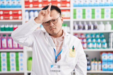 Photo for Chinese young man working at pharmacy drugstore making fun of people with fingers on forehead doing loser gesture mocking and insulting. - Royalty Free Image