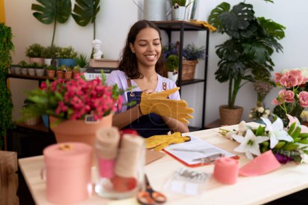 Téléchargez les photos : Jeune afro-américaine fleuriste souriant confiant portant des gants à la boutique de fleurs - en image libre de droit