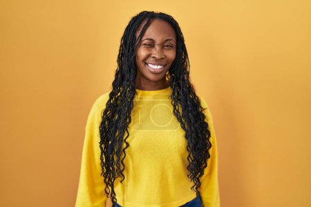 Téléchargez les photos : Femme africaine debout sur fond jaune clin d'oeil en regardant la caméra avec une expression sexy, gai et heureux visage. - en image libre de droit
