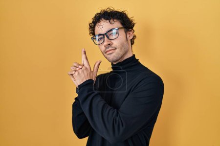 Photo for Hispanic man standing over yellow background holding symbolic gun with hand gesture, playing killing shooting weapons, angry face - Royalty Free Image