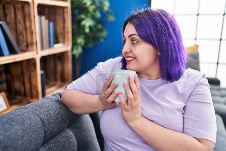 Téléchargez les photos : Jeune belle femme plus la taille de boire du café assis sur le canapé à la maison - en image libre de droit
