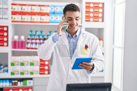 Photo for Young hispanic man pharmacist talking on smartphone using touchpad at pharmacy - Royalty Free Image