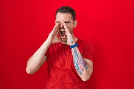 Téléchargez les photos : Jeune homme hispanique debout sur fond rouge criant en colère à haute voix avec les mains sur la bouche - en image libre de droit