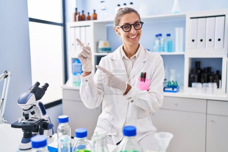 Photo for Young woman working at scientist laboratory smiling and looking at the camera pointing with two hands and fingers to the side. - Royalty Free Image