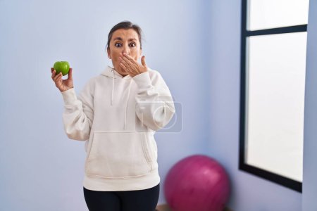 Téléchargez les photos : Femme hispanique du Moyen Âge portant des vêtements de sport tenant la pomme saine couvrant la bouche avec la main, choquée et effrayée par l'erreur. expression surprise - en image libre de droit