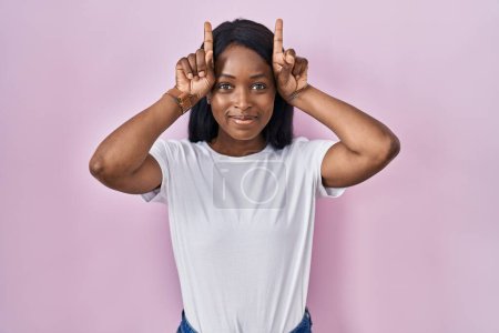 Téléchargez les photos : Africaine jeune femme portant casual t-shirt blanc faire geste drôle avec doigt sur la tête comme cornes de taureau - en image libre de droit