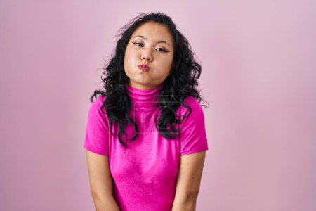Téléchargez les photos : Jeune femme asiatique debout sur fond rose joues gonflées avec un visage drôle. bouche gonflée d'air, expression folle. - en image libre de droit