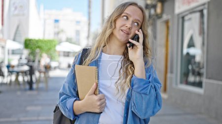 Photo for Young blonde woman student holding books talking on smartphone at street - Royalty Free Image