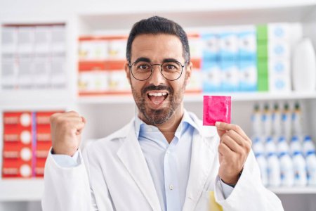 Téléchargez les photos : Homme hispanique avec barbe travaillant à la pharmacie pharmacie tenant préservatif criant fier, célébrant la victoire et le succès très excité avec les bras levés - en image libre de droit